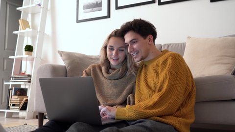 Happy young couple using laptop relaxing together at home. Smiling man and woman talking, looking at computer, surfing online website, having fun enjoying technology device in apartment together.