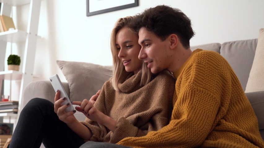 Young happy couple holding smartphone, looking at phone, enjoy playing mobile game, having fun using mobile cell app together sitting on sofa at home, betting or bidding in auction application. Royalty-Free Stock Footage #1066527412
