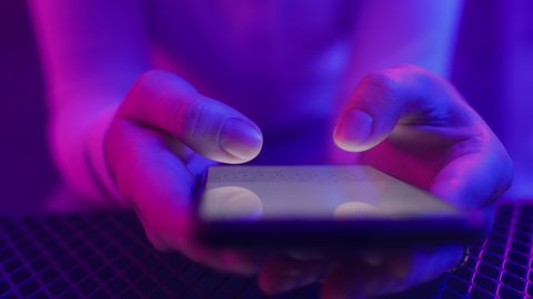 Use of mobile phone in shining neon light indoors. Creative vivid color of ultraviolet red and blue. Hands of person typing and messaging in chat closeup. Trendy neon room and colorful party concept