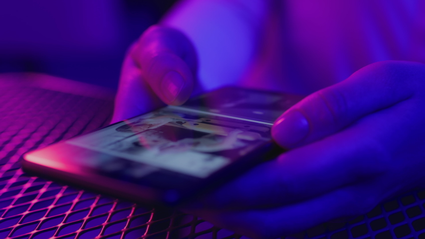 Use of mobile phone in shining neon light. Creative vivid color of ultraviolet red and blue. Hands of person scrolling up photos of instagram. Trendy social media photograph close-up at dark neon room