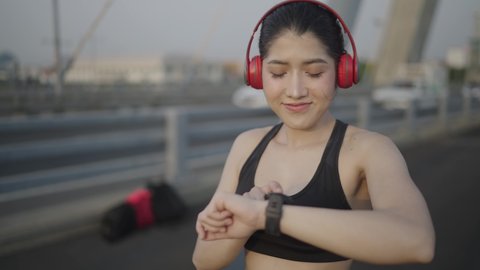 Slow motion video. A white Asian woman wearing a black sports bra. Black trousers And red headphones Viewing an exercise watch On a bridge across the river in the middle of the city
