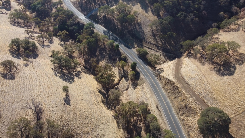 Traveling drone moving along isolated winding road lined with trees and golden hills
