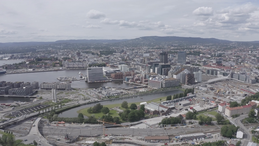 Oslo, Norway. The Oslo Opera House rises from the Oslofjord as a white marble-clad structure with sloped surfaces that extend to the waterline, creating a walkable public plaza that merges architecture with landscape. The aerial footage captures this contemporary landmark alongside the modern high-rise skyline of the Bjorvika district, with the full cityscape and surrounding hills visible in the background. Shot in flat logarithmic profile, the scene preserves wide tonal range for color grading. Daylight conditions with scattered clouds illuminate the waterfront, the angular opera house facade, and the dense urban fabric beyond. The camera moves forward at altitude, transitioning from a broad panoramic view to a closer perspective of the harbor and cultural quarter. The building
