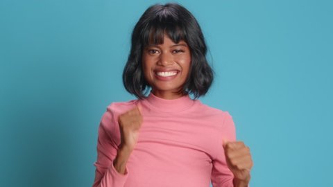 Happy dark skinned asian young woman makes triumph gesture says yes I did it makes victory dance glad to achieve great results dressed in pink polo neck poses against blue background. Success concept
