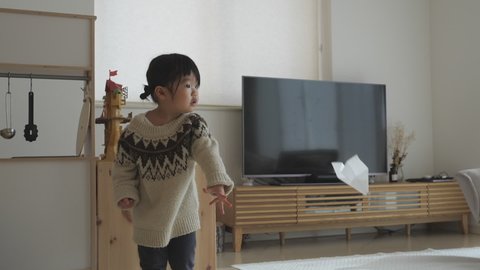 Funny Asian preschool toddler girl who can't fly a paper plane well in the living room at home. Large screen TV and playhouse kitchen background. Cute 2 or 3 years old child in sweater