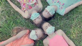 A group of girls lies on the grass in the park in medical masks and pull their hands up. Virus protection. 4K video.