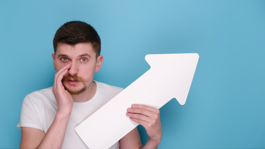 Senior young bearded man with white paper arrow holding hand on mouth telling secret or rumor, whispering it to camera, dressed in t-shirt, models over blue background studio with copy space | Shutterstock HD Video #1066557901