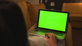 Young woman using laptop with green screen, lying on the couch sofa at the home. Back view. Chroma key