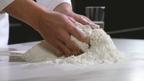 Professional baker works in kitchen of bakery shop. cook carefully mixes white wheat flour with chicken egg on table with his hands. Kneading dough. Preparation of dough for baking bread and pizza.