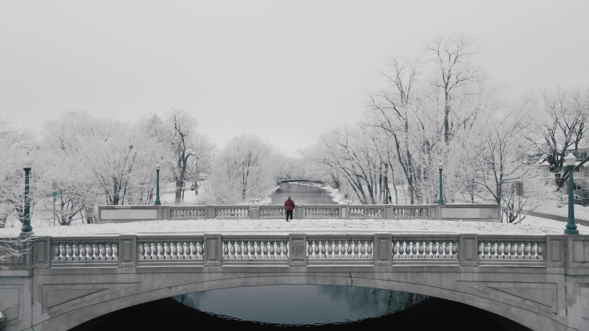 Aerial video swooping over a person standing on a picturesque stone bridge and revealing a winter river lined with white snowy trees.