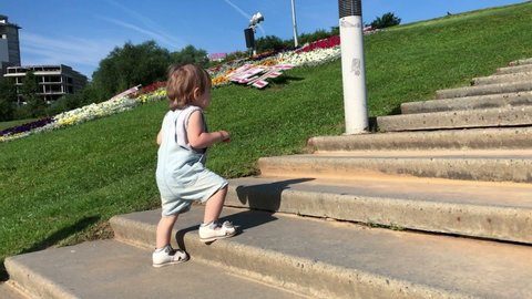 Toddler Caucasian girl wearing overall climbing on stone stairs outdoor, wide cement stairway up to hill