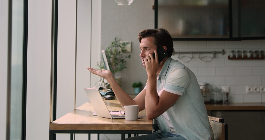 Relaxed young male freelancer or businessman sitting at table with laptop in modern kitchen, discussing project details with colleagues by mobile phone call, drinking coffee, casual distant workday. Royalty-Free Stock Footage #1066586218