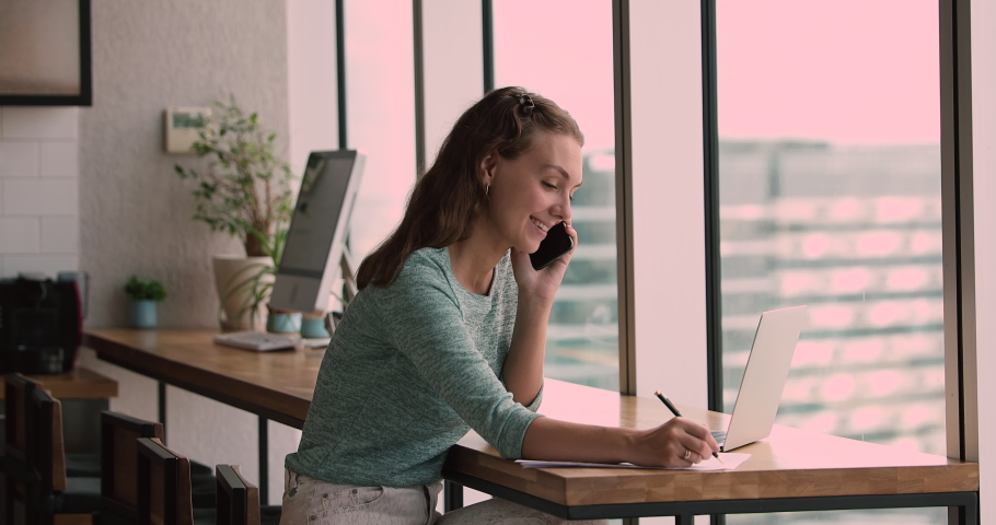 Happy millennial woman holding cellphone conversation, booking hotel or tickets distantly, making order in internet store, asking information about job vacancy, writing notes on paper at home. Royalty-Free Stock Footage #1066586389