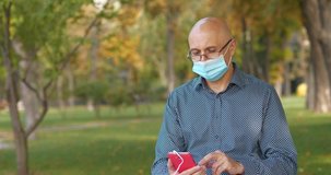Bald senior man in glasses and medical protective face mask sits on the bench in city park, puts on earphones and listen to music on his smartphone. Cinema 4K 60fps video