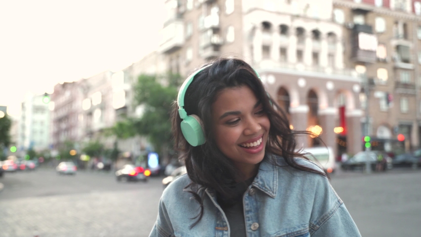 Happy cool African American woman wearing headphones dancing alone on street. Smiling young mixed race lady hipster listening music standing in city outdoors, feeling free and funky. | Shutterstock HD Video #1066593487
