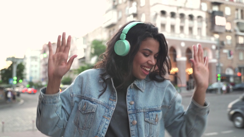 Happy cool African American woman wearing headphones dancing alone on street. Smiling young mixed race lady hipster listening music standing in city outdoors, feeling free and funky.