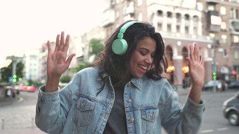 Happy cool African American woman wearing headphones dancing alone on street. Smiling young mixed race lady hipster listening music standing in city outdoors, feeling free and funky.