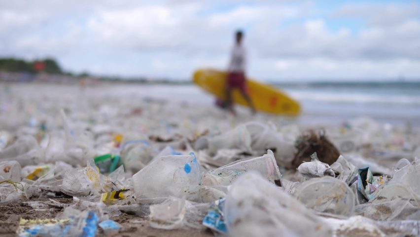 Close up of plastic pollution on the beach. Man with the surfboard in the background  | Shutterstock HD Video #1066599910