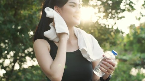 Young pretty Asian woman in black tank top standing wiping sweat with white towel draped around her neck while smiling looking at the water bottle in hand with blurred background of trees and sunlight