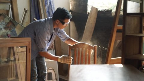 Young Asian male carpenter in safety goggles and gloves rubbing and blowing off dust on the backrest of a finished wooden chair and cleaning dust from his gloves by clapping hands in the wood workshop