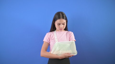Portrait of young woman with an injured arm in a sling over blue background in studio, insurance and healthcare concept