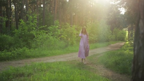 A beautiful woman smiles as she walks along a path surrounded by foliage and trees. Front view of a woman in a dress walking in a summer Park. Slow motion