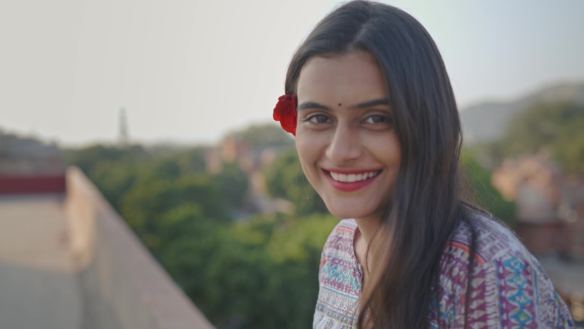 cheerful young beautiful Indian female in traditional dress wearing red rose in hair looking at camera, raising eyebrows and laughing on top of a buildings terrace in a daylight Royalty-Free Stock Footage #1066612792