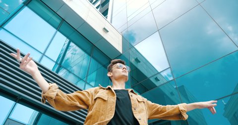 Closeup stylish attractive guy teen man wears fashionable jacket and sunglasses, dances on street modern movements, spins by hand, moves pliable flexibly, has fun outdoor near glass urban building