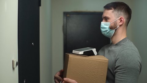 Side view of courier in medical mask coming to door with parcel and contactless payment terminal, rings doorbell to meet customer and hand over online order. Delivery man waiting client in entryway.