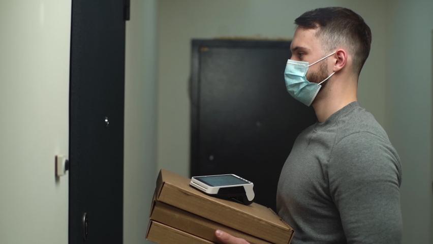 Courier in medical mask coming to door with boxes with hot pizza and contactless payment terminal, rings doorbell to meet customer and hand over online order. Delivery man waiting client in entryway. | Shutterstock HD Video #1066628341
