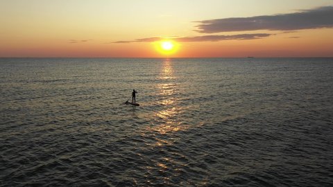 Young woman on a stand up paddle board exercising at sunset. Stand Up Paddle Board Girl Silhouette on Water. Active sporty girl standing on inflatable SUP, paddling at amazing golden sunset, sunrise