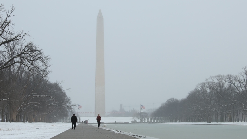 Washington, DC - United States - February 1 2021: A runner and a masked pedestrian near the Lincoln Memorial Reflecting Pool on a snowy day. The Washington Monument is seen in the background.
