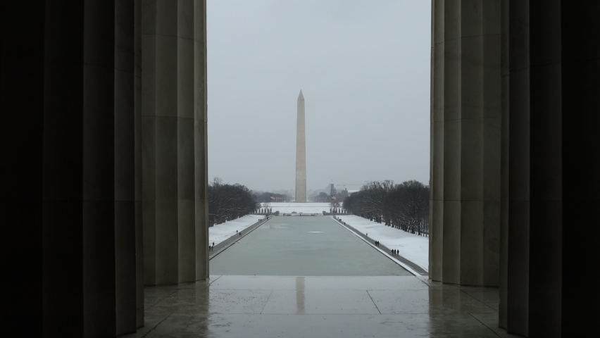 The Washington Monument and Reflecting Pool as seen from inside the Lincoln Memorial on a snowy winter day in Washington, D.C. Architectural columns frame the shot on both sides.