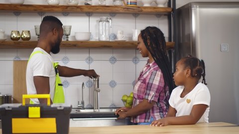 African plumber repairing running water for mother and daughter. Side view of afro-american handyman showing installed faucet to young woman and little girl in kitchen