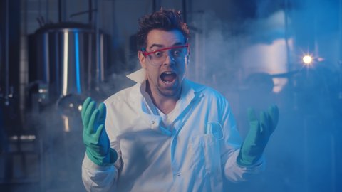 Portrait of crazy scientist laughing at camera in large smoky laboratory. Man chemist in lab coat, gloves and glasses laughing standing in industrial lab