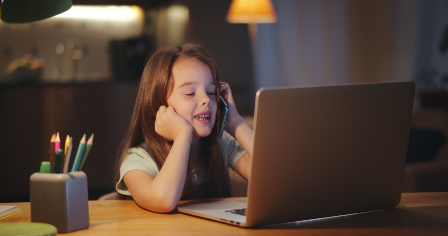 Little girl talking on smartphone and watching cartoons on laptop sitting at table in dark living room. Portrait of preteen kid having phone call and studying on computer late in evening | Shutterstock HD Video #1066634647