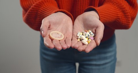 Close up of woman holding birth control pills and condom isolated on grey background. Cropped shot of female with contraception medication and condom. Safe sex concept