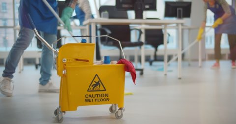 Cropped shot of janitors with mop cleaning modern office floor. Focus on yellow bucket with equipment and staff wiping floor and furniture in office space