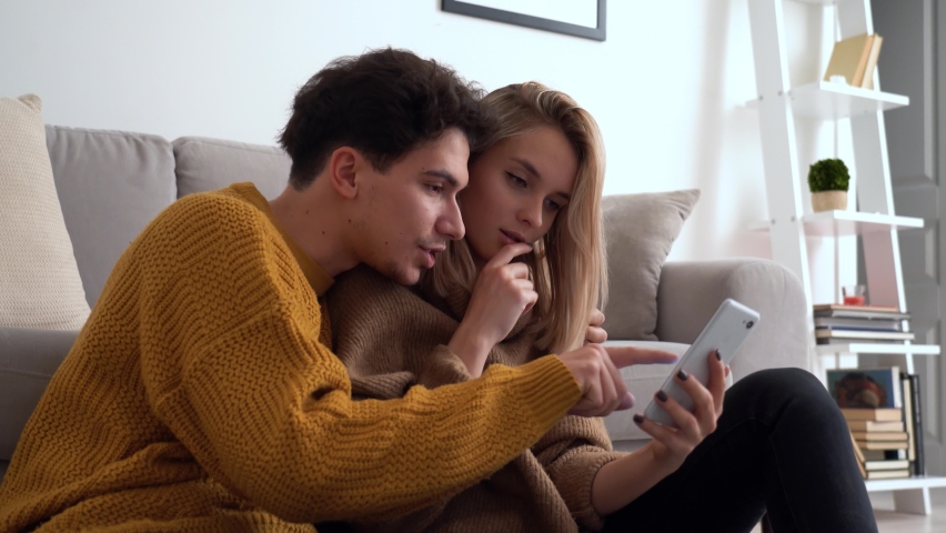 Young happy couple having fun holding smartphone using mobile phone at home, laughing, playing game, doing online shopping in ecommerce app together, checking social media applications together. Royalty-Free Stock Footage #1066636111