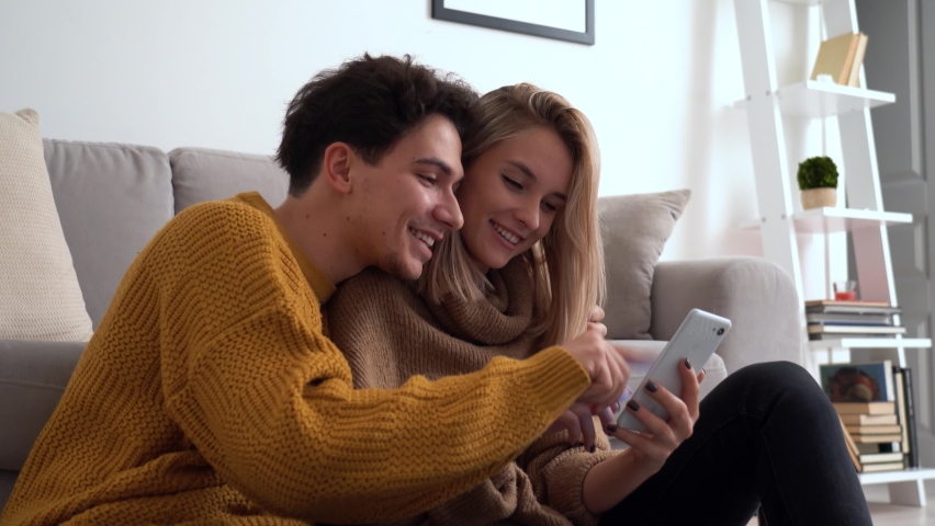 Young happy couple having fun holding smartphone using mobile phone at home, laughing, playing game, doing online shopping in ecommerce app together, checking social media applications together. | Shutterstock HD Video #1066636111