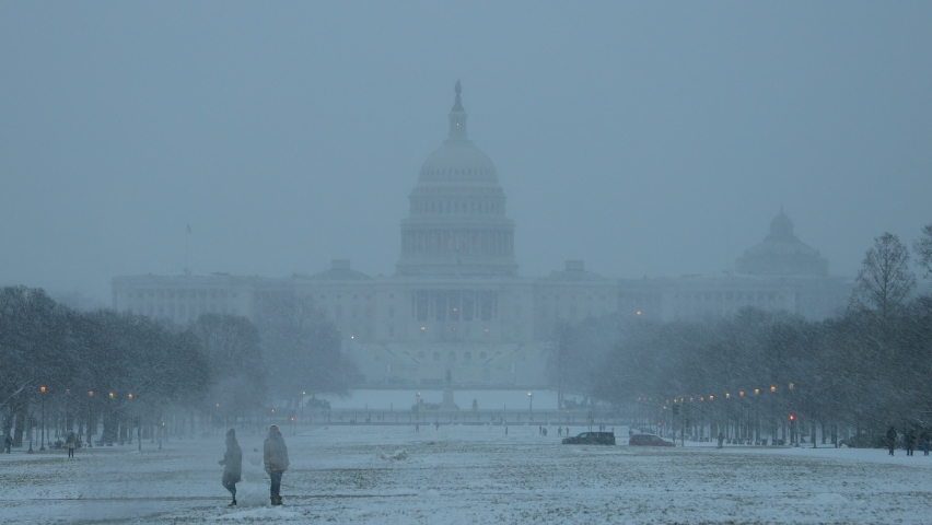 Washington, DC - United States - February 1 2021: The west front of the U.S. Capitol building and National Mall seen during a winter snowstorm. A snowman is seen in the foreground as snowflakes fall.