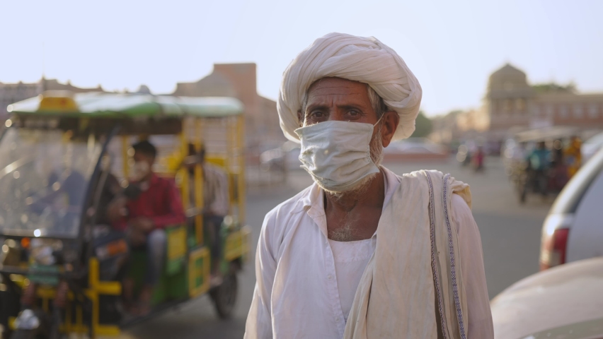 Close shot of an old Indian male villager in traditional attire wearing face protective mask and standing next to a busy road in a market place while looking at the camera amid Corona virus epidemic | Shutterstock HD Video #1066638247