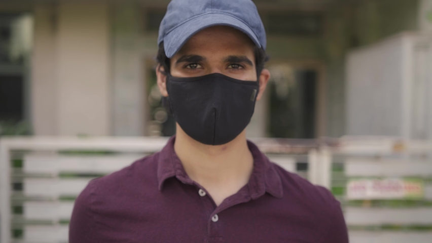 Track-in shot of a young Indian Asian delivery boy with a protective mask on face and parcel in hand standing in front of a house looking at camera during covid 19 epidemic Royalty-Free Stock Footage #1066638250