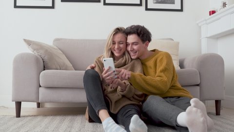 Young happy couple having fun using smartphone at home. Girl and guy talking, laughing, looking at mobile phone buying retail products shopping online, ordering delivery, checking social media apps.