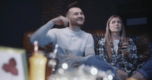 Young couple sitting on leather sofa watching TV musical show at home in evening. Guy is dancing funny while watching music video, girl surprised and shocked watching her boyfriend crazy dance