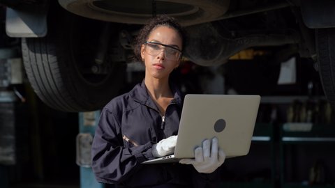 african american mechanic woman using laptop computer  checking list to undercarriage of car in workshop at auto car repair service center with lift . car engineer female inspection vehicle details