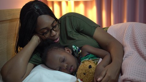 african american mother singing lullaby for little daughter before going to Sleep on bed at home. black mom story telling to girl in bedroom. sleeping child in parent embrace . kid hug Teddy bear 