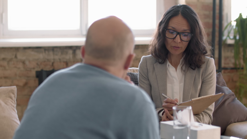 Tracking shot of female psychotherapist in glasses writing notes on clipboard and talking to emotional male client during therapy session in her private practice | Shutterstock HD Video #1066644802