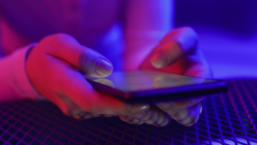Use of mobile phone in shining neon light of night event indoors. Creative art color of ultraviolet red and blue. Hands of stylish woman typing and messaging in chat close-up in vivid multicolour club | Shutterstock HD Video #1066645453