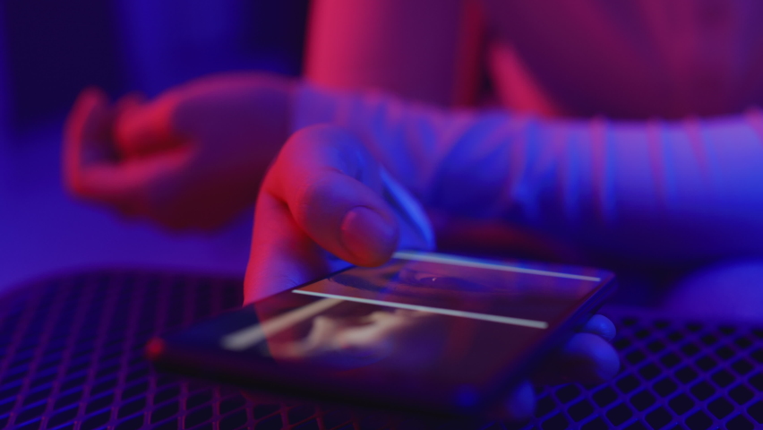 Use of mobile phone in shining neon light. Creative vivid color of ultraviolet red and blue. Hands of person scrolling up photos of instagram. Stylish social media photograph close-up at neon art room